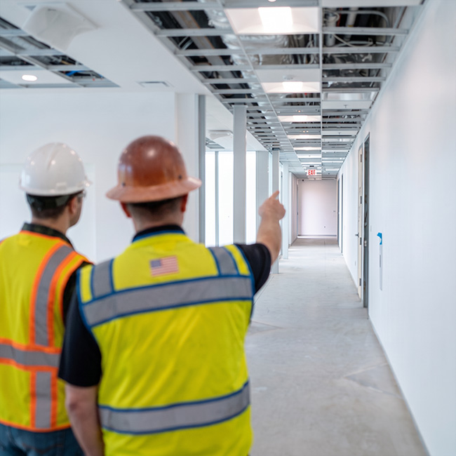 Two construction workers in hard hats and safety vests view an unfinished hallway with exposed ceiling and white walls.