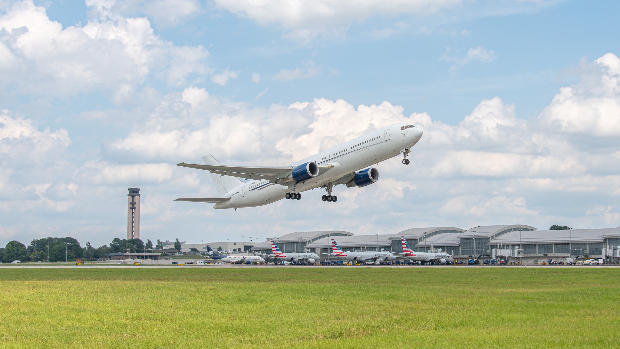 A large white airplane takes off from an airport runway with several American Airlines planes in the background. A control tower is visible against a partly cloudy sky.
