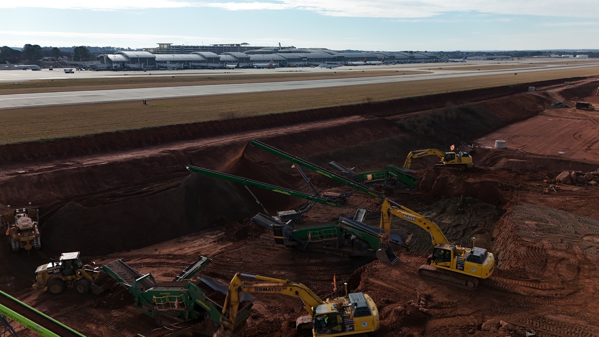 Runway 5L-23R construction site with yellow excavators and green screening equipment working on brown earth, with RDU's current runway and Terminal 2 in background.
