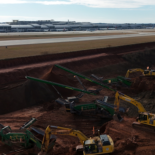 Runway 5L-23R construction site with yellow excavators and green screening equipment working on brown earth, with RDU's current runway and Terminal 2 in background.