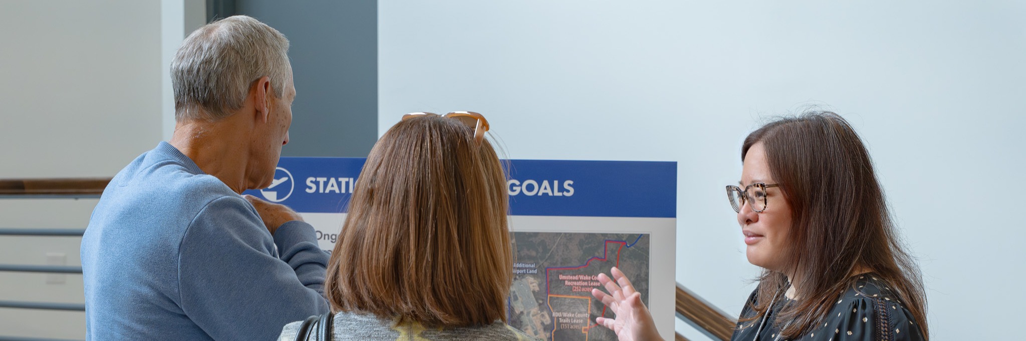 Woman in glasses presenting to two people in front of a blue banner displaying station goals with a map.