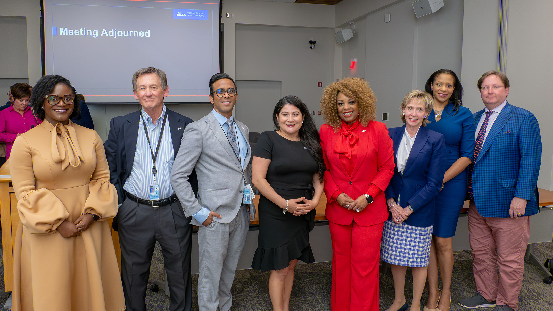 Group of professionally dressed people standing in a meeting room with a projection screen displaying Meeting Adjourned.