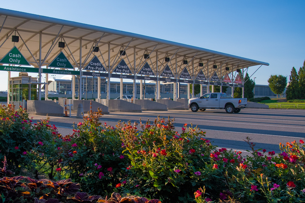 Modern toll plaza with multiple lanes under a blue canopy, white truck passing through, colorful flowers in foreground.