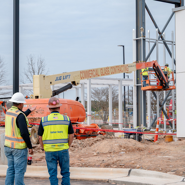 Construction workers in safety vests observe an orange lift and crane at a building site with steel framework.
