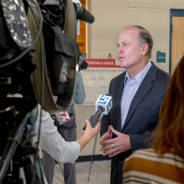 A person is speaking to reporters and camera crews in a room with beige walls. Several microphones, including one with the logo "KWTX," are directed toward the speaker.