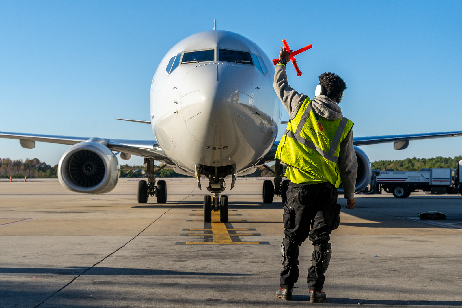 Ground crew worker in yellow safety vest guides white commercial airplane on tarmac with red signal wands under clear blue sky.