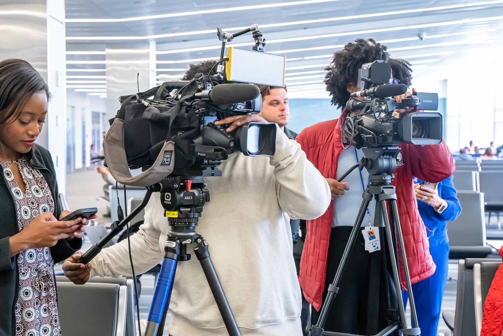 News crew filming in a bright modern building with two camera operators and a woman holding a microphone.