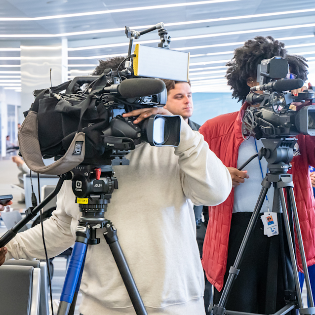 News crew filming in a bright modern building with two camera operators and a woman holding a microphone.
