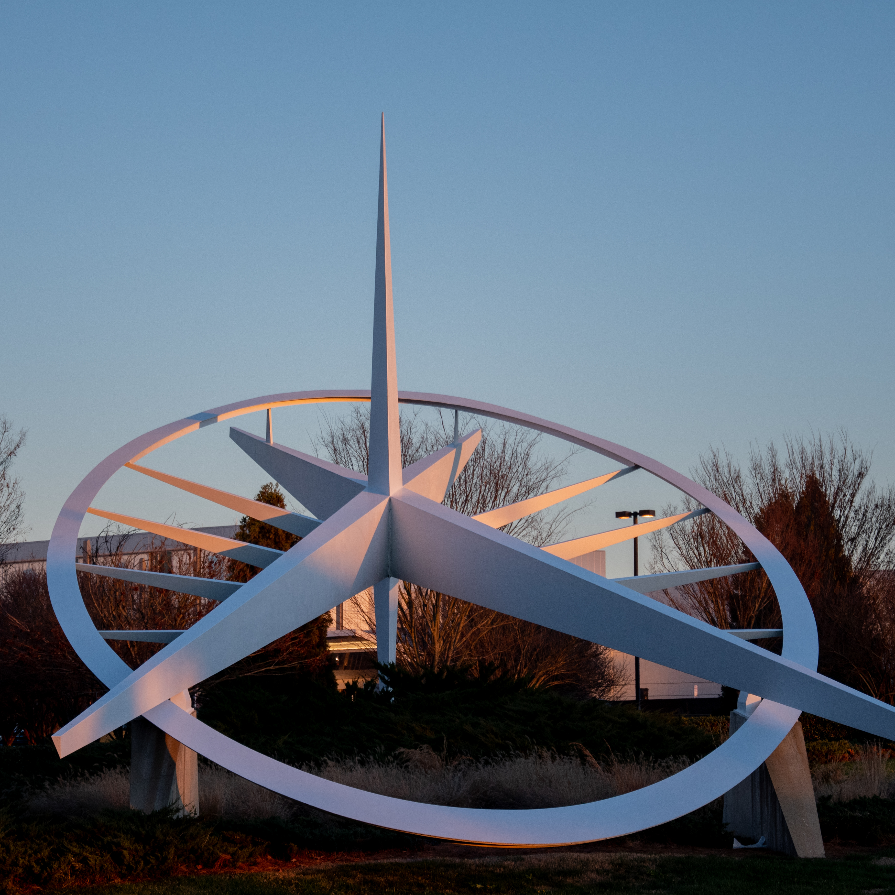 White modern sundial sculpture with curved metal bands and central spire against blue sky at dusk with bare trees behind.
