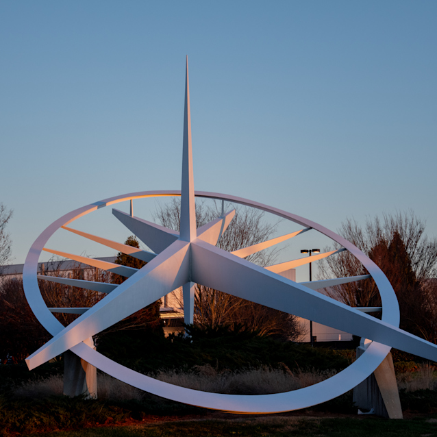 White modern sundial sculpture with curved metal bands and central spire against blue sky at dusk with bare trees behind.