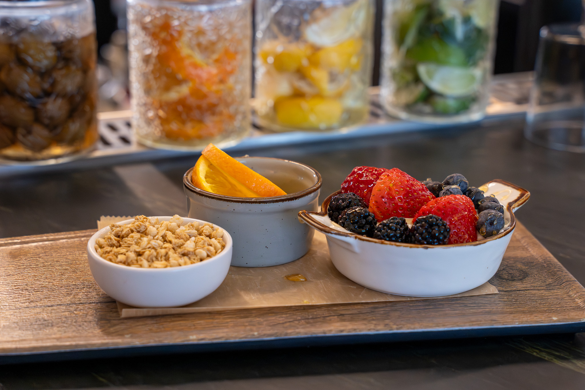 A wooden tray displays a small bowl of granola, a bowl of mixed berries including strawberries, blackberries, and blueberries, and a mug with orange slices. In the background, glass jars containing various preserved fruits or vegetables are lined up.

