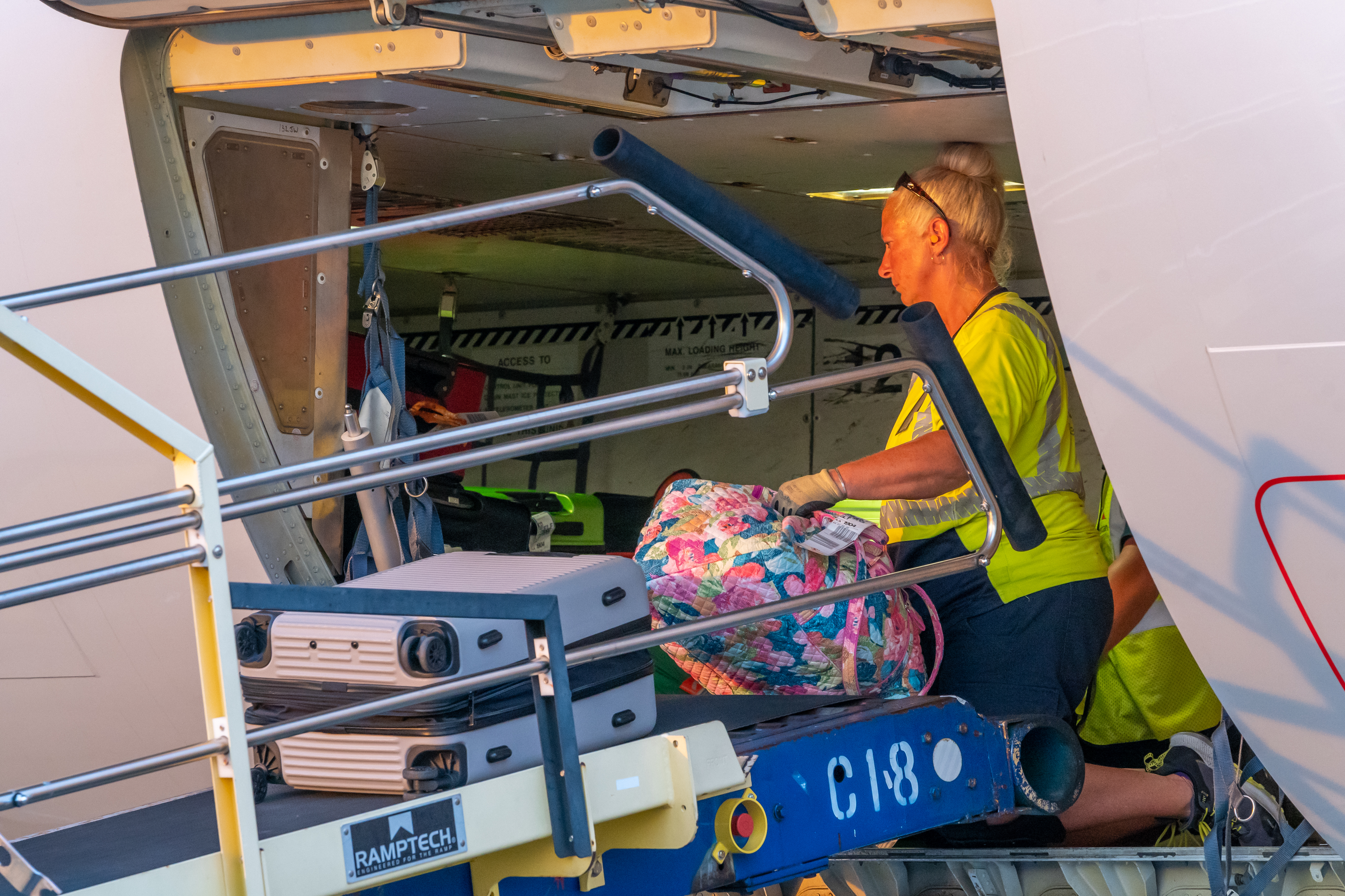 Female baggage handler in yellow safety vest loading colorful luggage onto aircraft cargo hold via conveyor lift.
