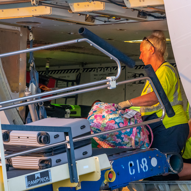 Female baggage handler in yellow safety vest loading colorful luggage onto aircraft cargo hold via conveyor lift.