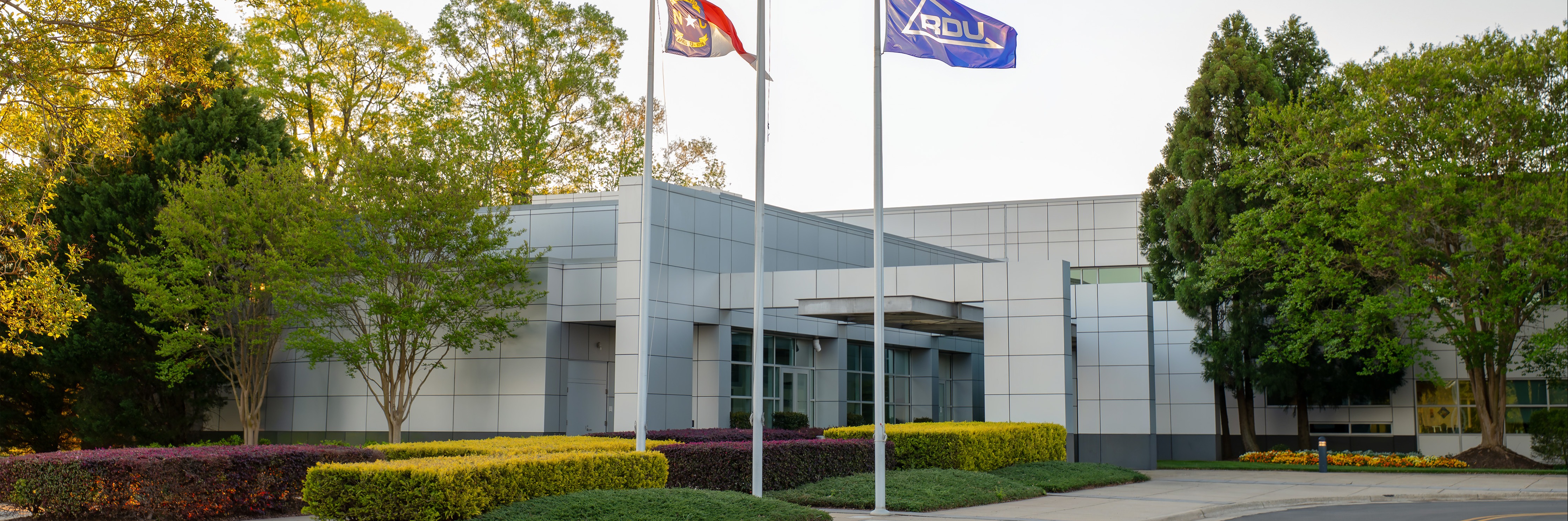 Three flags flying in front of a modern white building with green landscaping, trees, and a plane overhead in the sky.