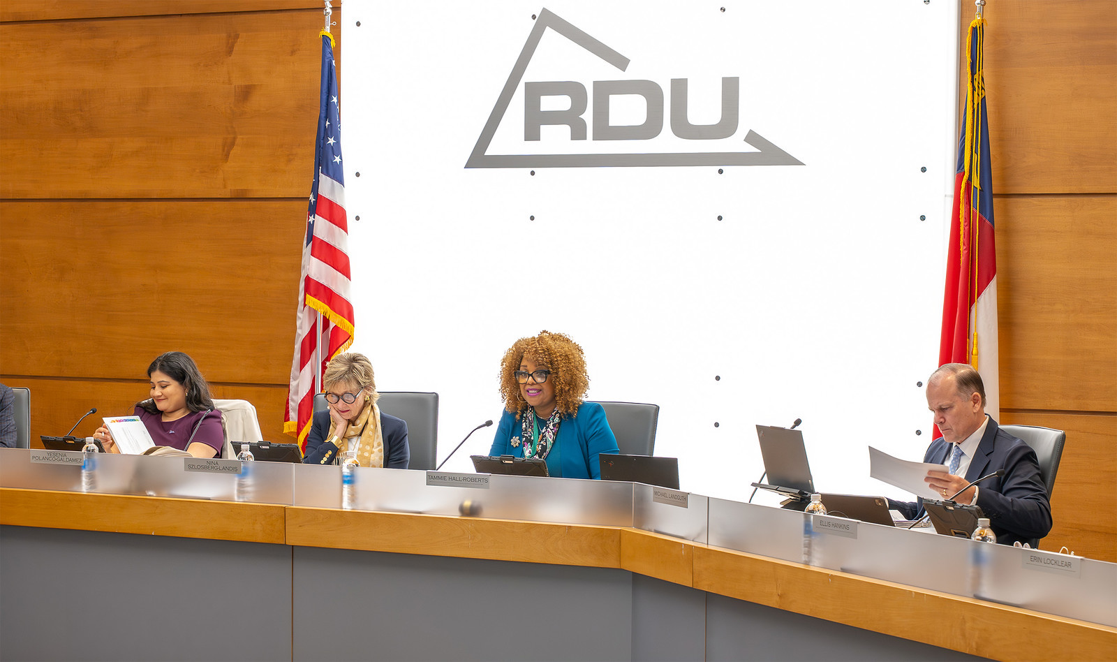 Four board members seated at a wooden table with laptops and microphones, flanked by U.S. and state flags, beneath RDU logo.