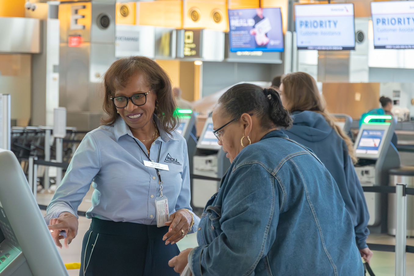 Airport employee in blue shirt and glasses assists customer in denim jacket at service counter with digital screens above.
