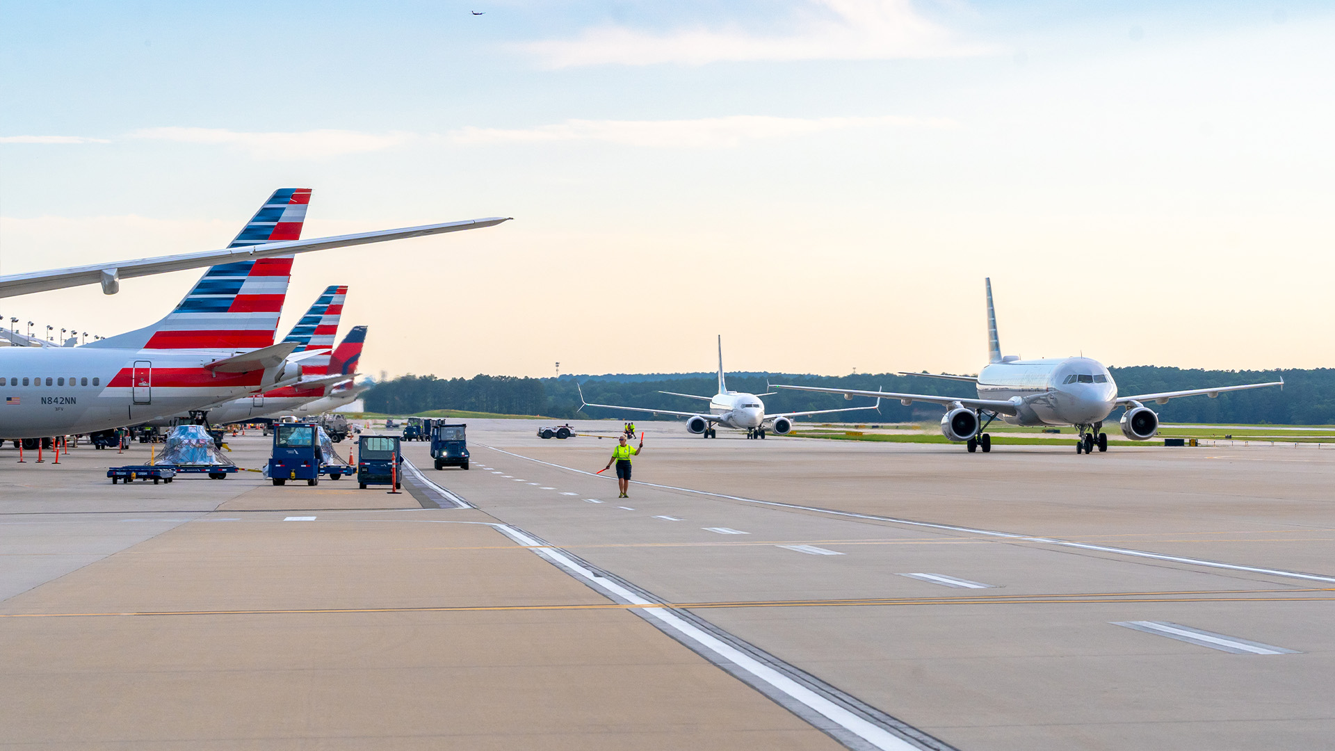 A runway scene with multiple airplanes from Delta Airlines and American Airlines. The American planes display their distinctive tail graphics with a Delta tail visible in the far background.
