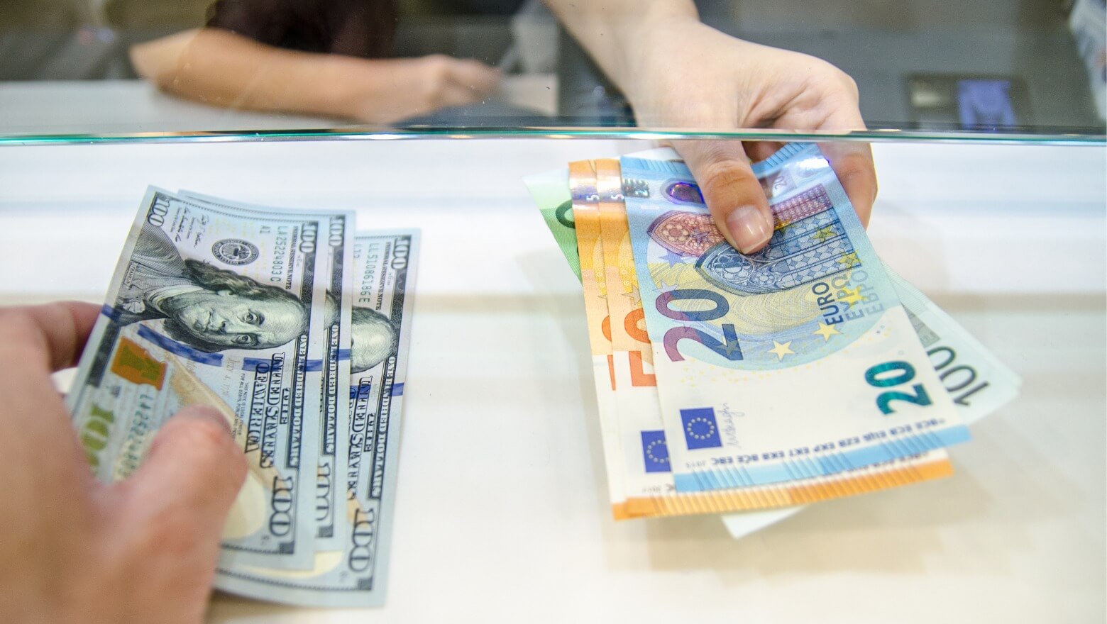 Two people exchanging currency through a glass partition, one holding US dollars and the other holding Euro bills.
