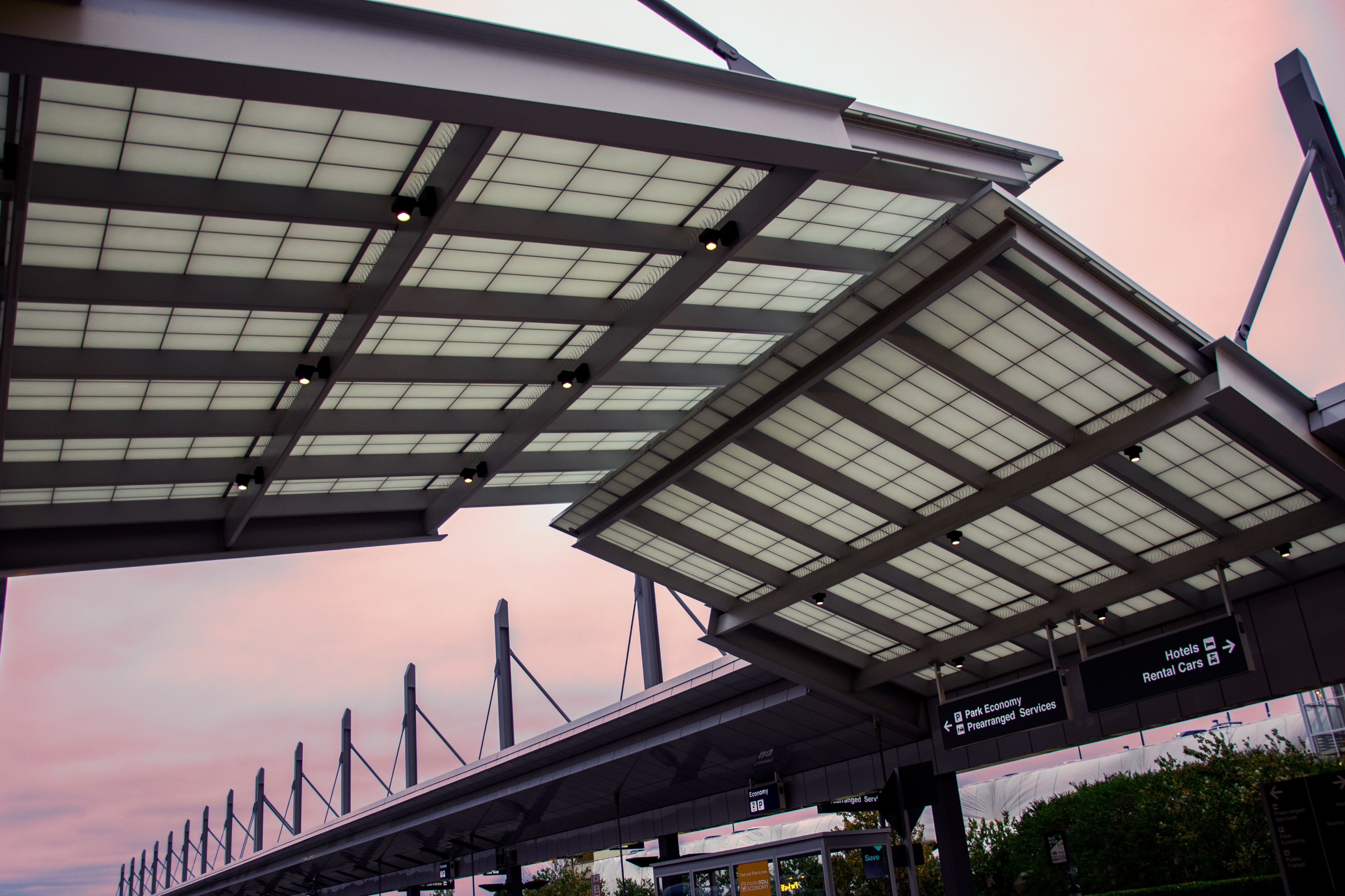 Modern glass and steel canopy structure with translucent panels and dark beams against a pink sunset sky at an airport.