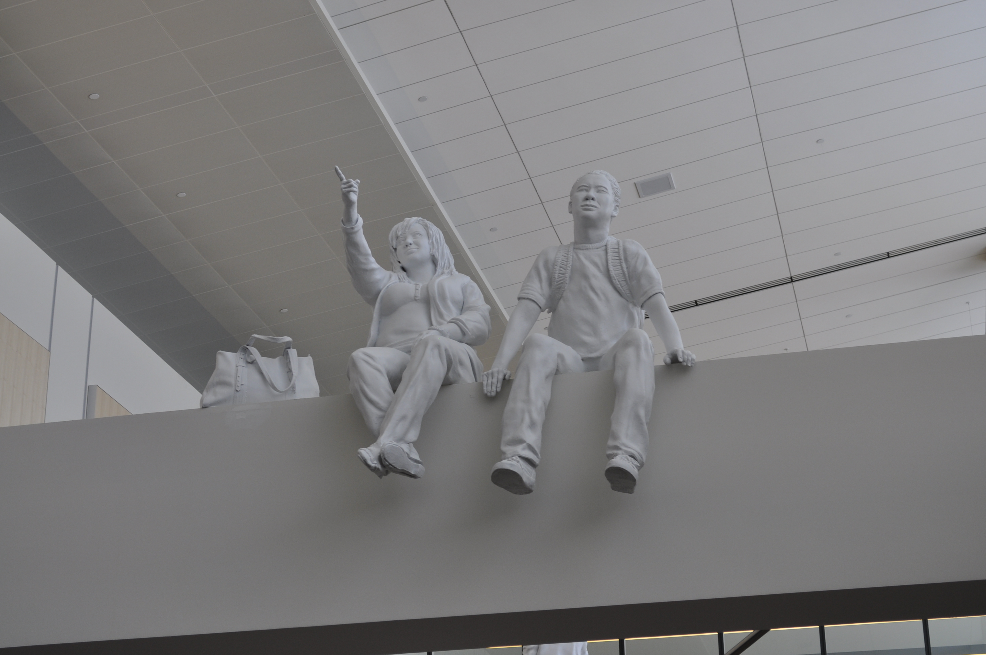 White sculpture of two children sitting on a ledge, one pointing upward, displayed in a modern building interior.
