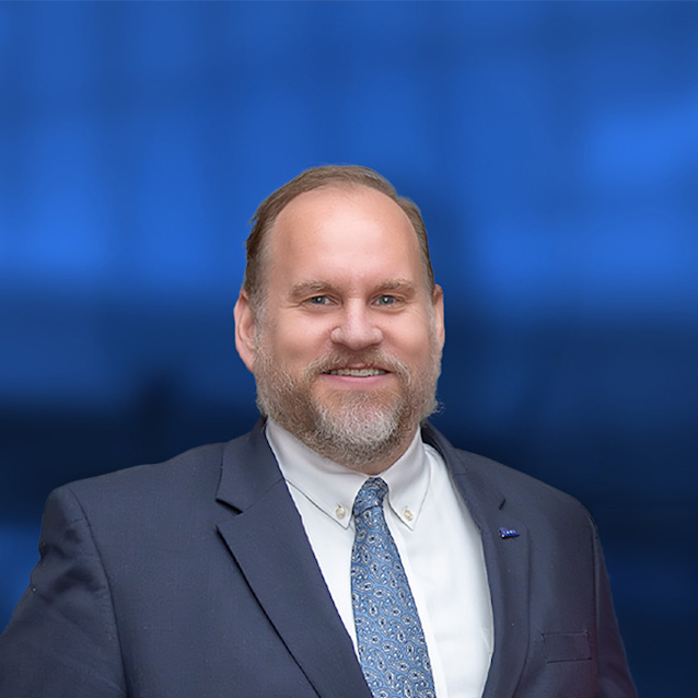 Professional headshot of a smiling man with a beard wearing a dark suit, white shirt, and blue patterned tie against a blue background.