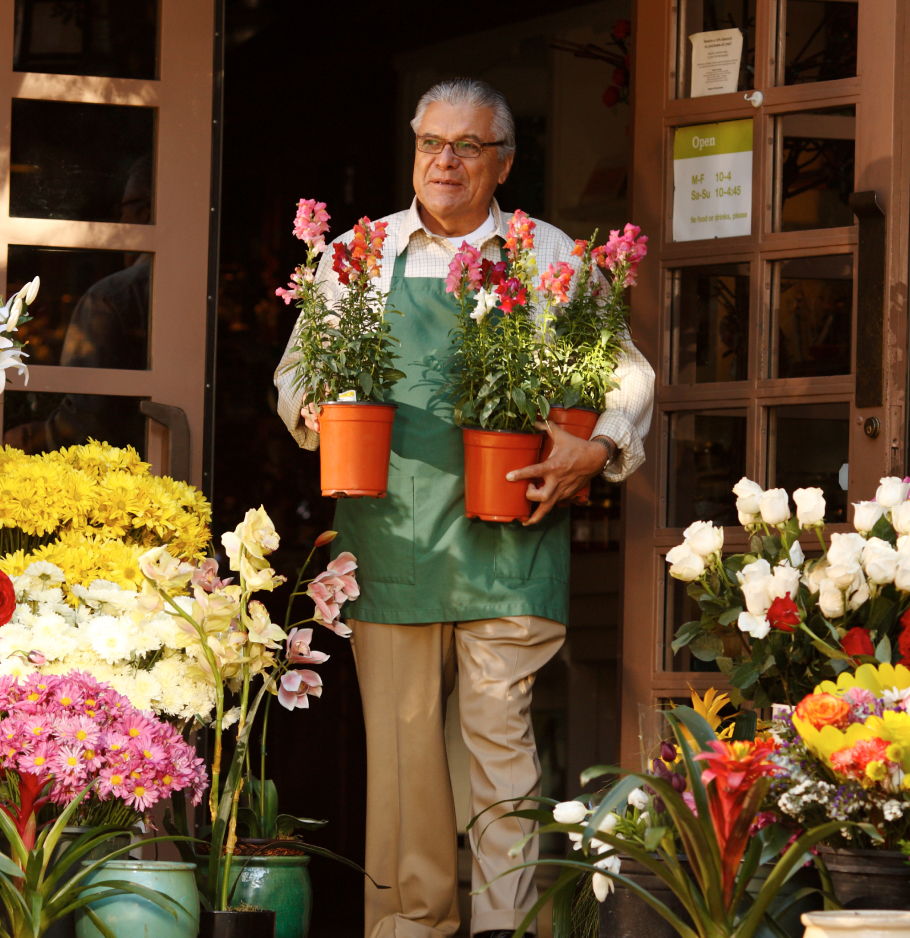 A business owner standing in the doorway of their business holding two pots of plants