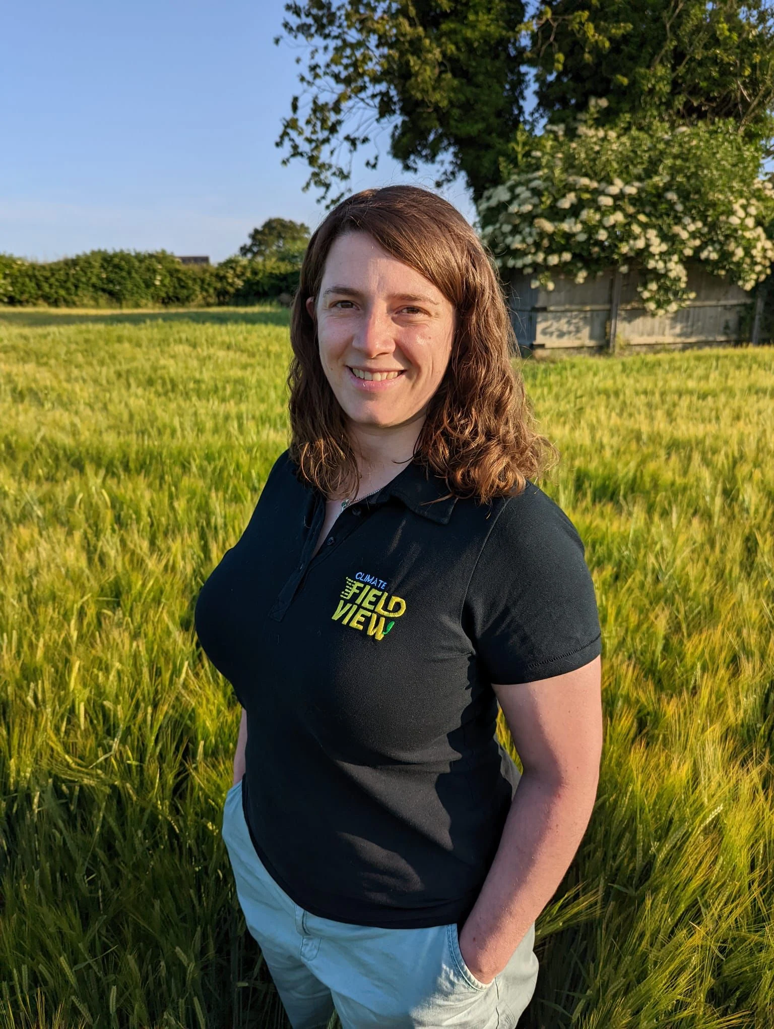 Woman smiling, with long brown hair and black top is stood in a crop field