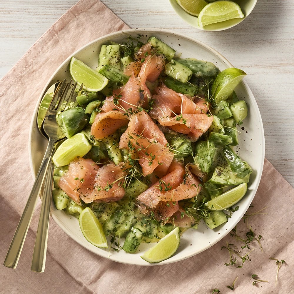 Gravad lax med asiatisk hovmästarsås, gurka och limeklyftor upplagda på en tallrik med bestick bredvid.
