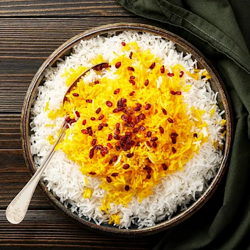 En skål med vit och saffransfärgad ris, toppad med röda bär, på ett träbord. En sked ligger lutad mot skålen.