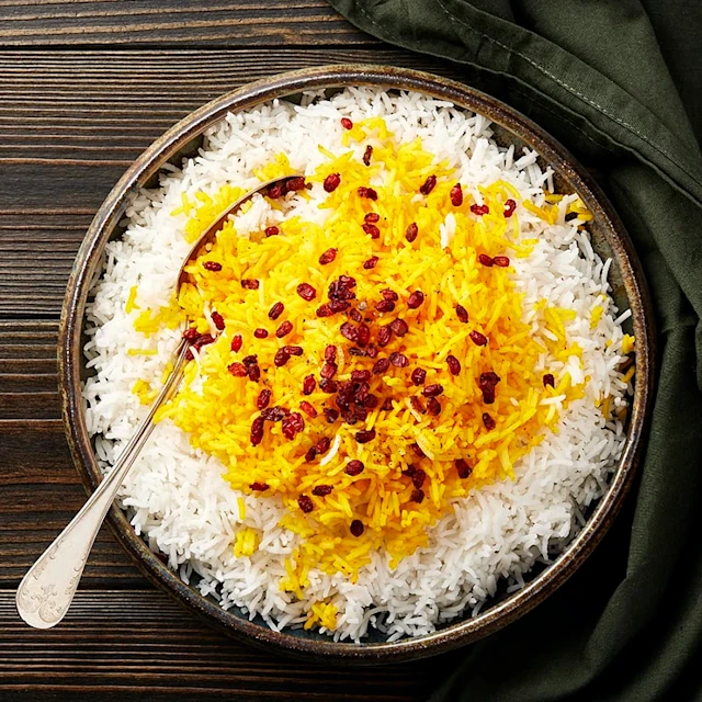 En skål med vit och saffransfärgad ris, toppad med röda bär, på ett träbord. En sked ligger lutad mot skålen.