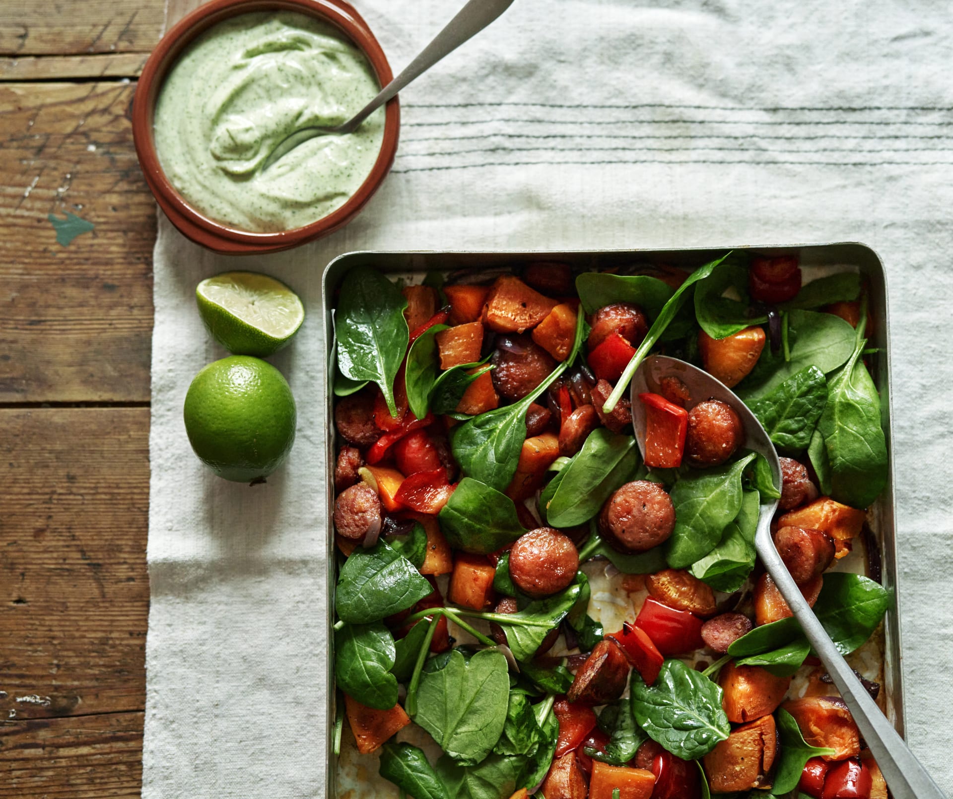 Sötpotatis och korv på plåt med färsk spenat, serverad med koriandermajonnäs och limehalvor bredvid.