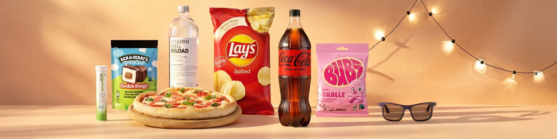 Snacks och dryck uppställda på bord: glass Ben & Jerry’s Cookie Dough, brustabletter, pizza, Vitamin Well Reload, chips Lay’s Salted, Coca‑Cola Zero Sugar och godis BUBS Skalle. Klicka för att handla.