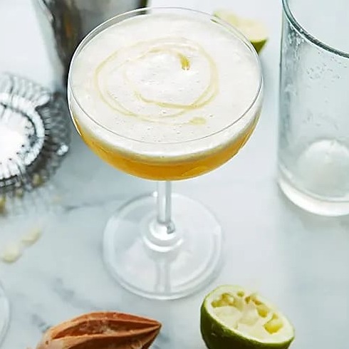 Två cocktailglas med skummig gul drink, pressad limehalva och träslev på marmorbord.