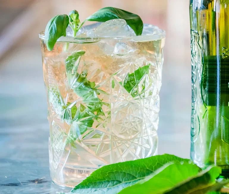 Drink med is och gröna örter i mönstrat glas, bredvid gröna blad och flaska.