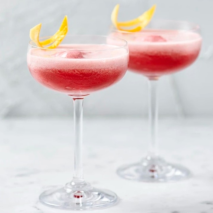 Rosa drinkar med skum i cocktailglas, dekorerade med citronskal på marmorbord.