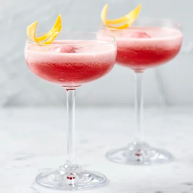 Rosa drinkar med skum i cocktailglas, dekorerade med citronskal på marmorbord.