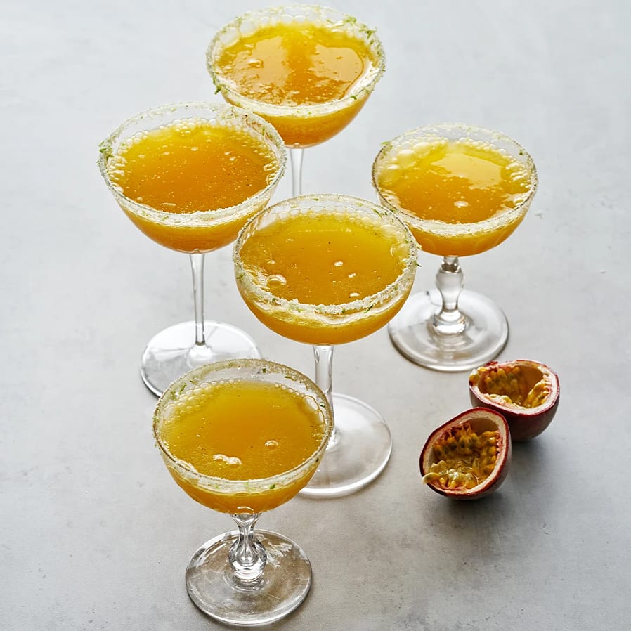 Fem cocktailglas fyllda med gul passionfruktsdrink, med socker på kanten, bredvid delad passionfrukt.
