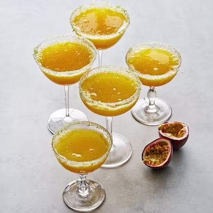 Fem cocktailglas fyllda med gul passionfruktsdrink, med socker på kanten, bredvid delad passionfrukt.