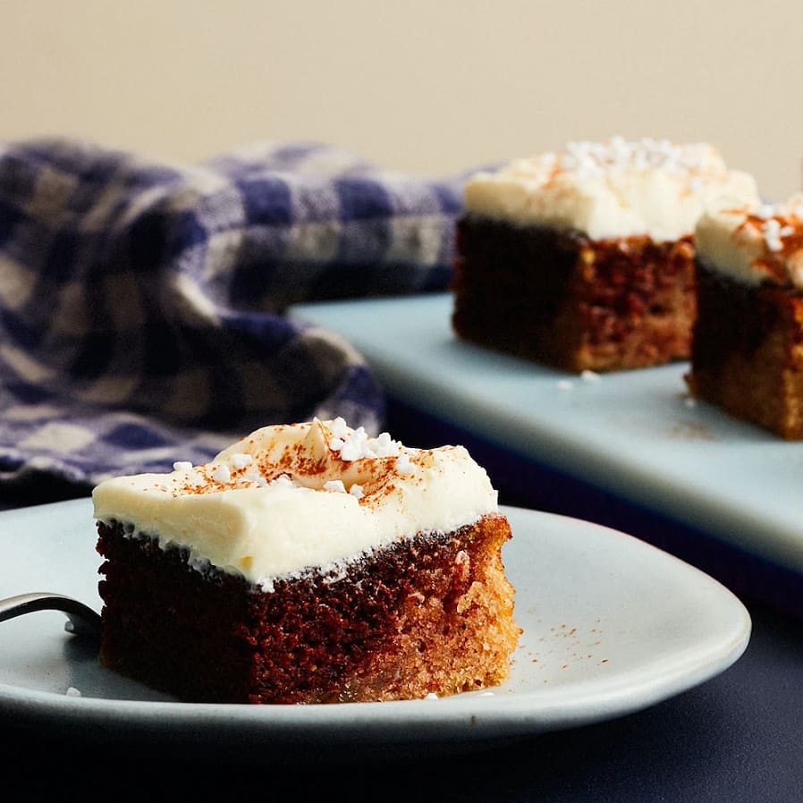 En bit kaka med vit grädde och chokladbotten, serverad på en blå tallrik med en sked. I bakgrunden syns fler kakedelar.