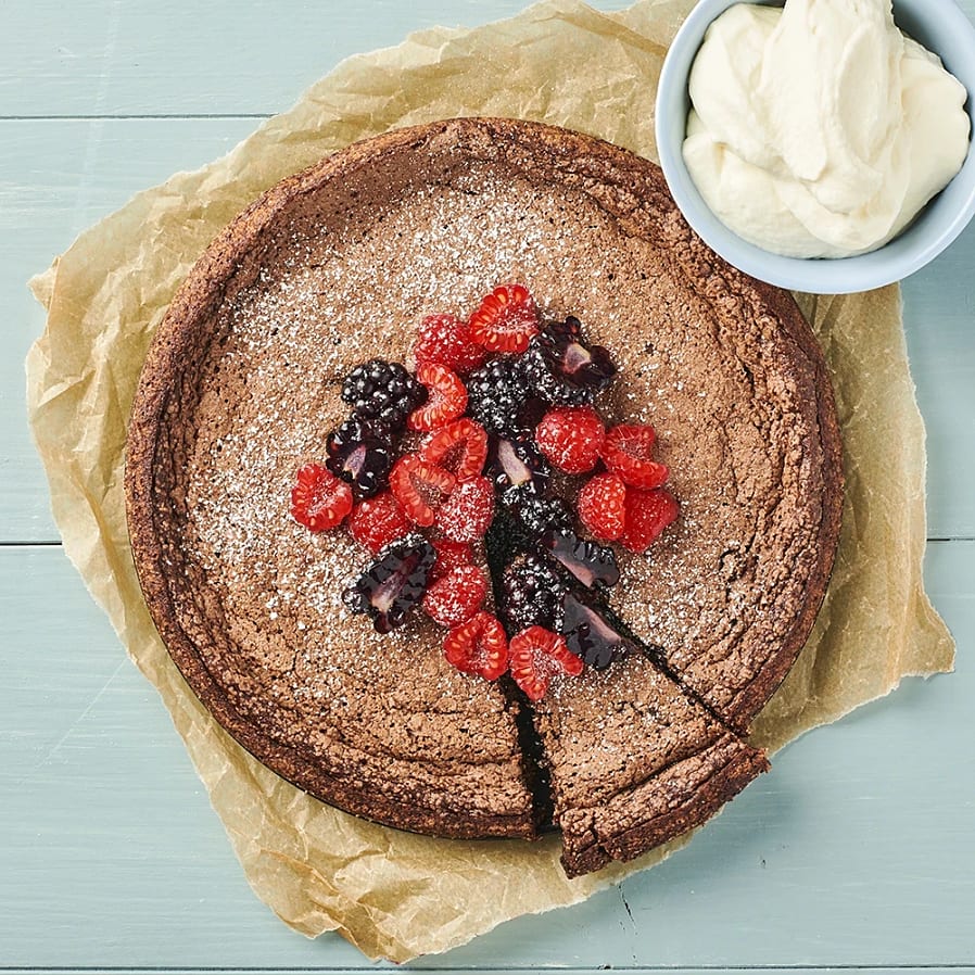 En rund kladdkaka med björnbär och hallon som ligger i mitten. Kakan ligger på en bit bakplåtspapper och i bakgrunden syns ett blått bord. I sida av kladdkakan står en liten skål med vispad grädde 