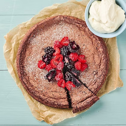 En rund kladdkaka med björnbär och hallon som ligger i mitten. Kakan ligger på en bit bakplåtspapper och i bakgrunden syns ett blått bord. I sida av kladdkakan står en liten skål med vispad grädde