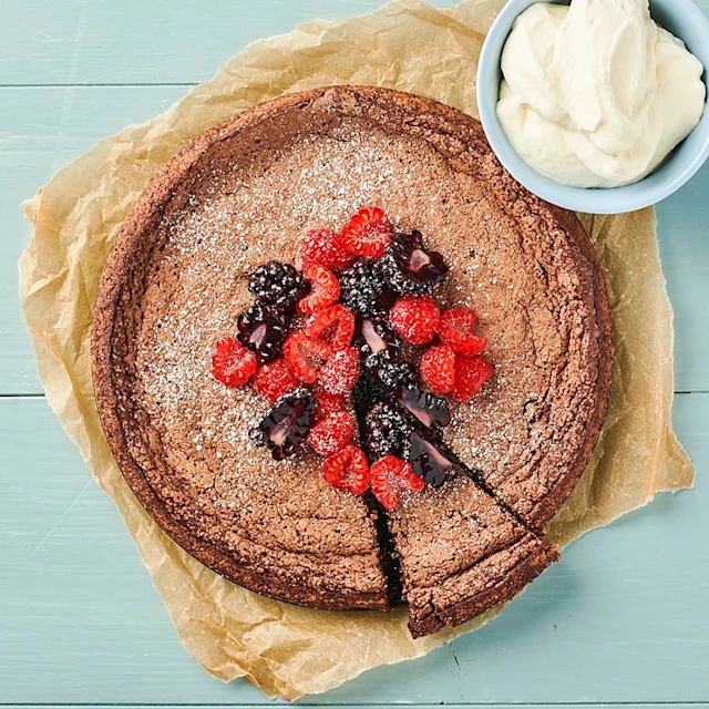 En rund kladdkaka med björnbär och hallon som ligger i mitten. Kakan ligger på en bit bakplåtspapper och i bakgrunden syns ett blått bord. I sida av kladdkakan står en liten skål med vispad grädde