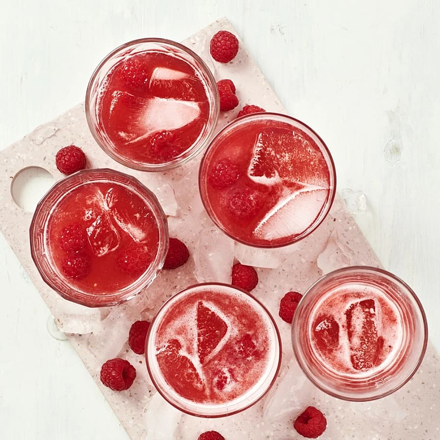 Fem glas med röd hallondryck med is och färska hallon på ett ljus underlägg med isbitar.