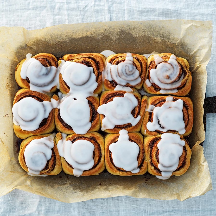 Kanelbullar med glasyr i en ugnsform, arrangerade i fyra rader och tre kolumner, på ett bakplåtspapper.