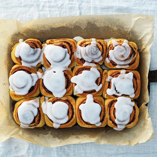 Kanelbullar med glasyr i en ugnsform, arrangerade i fyra rader och tre kolumner, på ett bakplåtspapper.