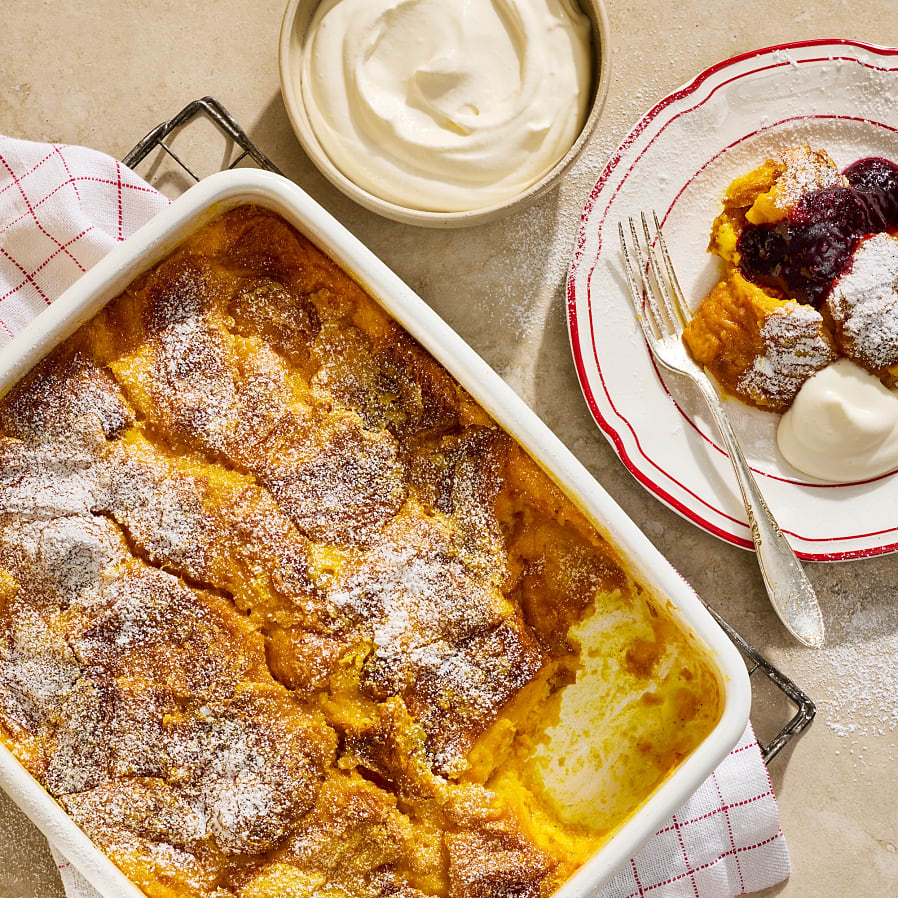 En gratängform med saffranspudding pudrad med florsocker, en bit tagen som ligger på en assiett toppad med yoghurtgrädde och sylt.