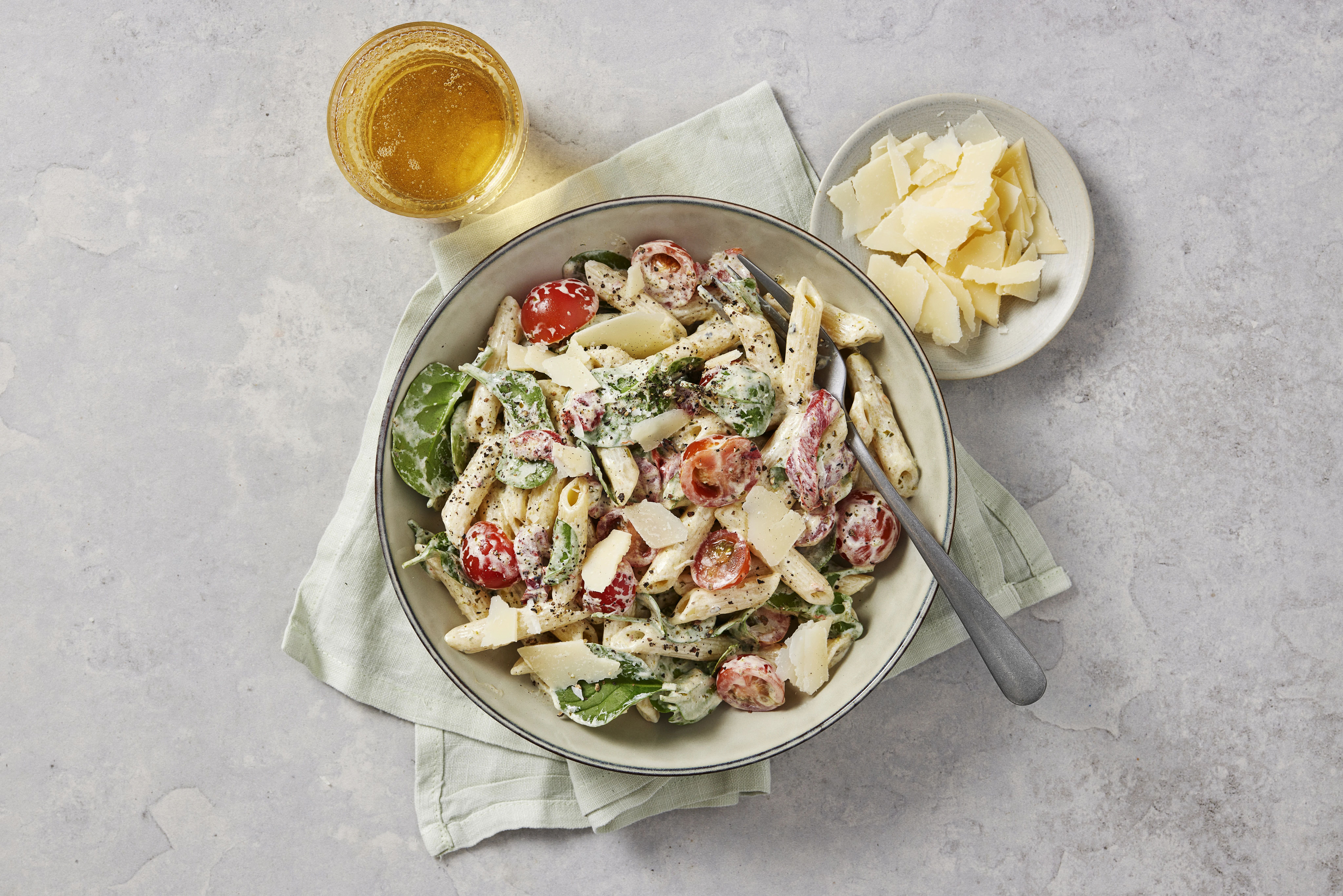 Krämig pastasallad med penne, babyspenat, körsbärstomater och parmesanflarn, serverad i en skål med dryck och extra parmesan bredvid.
