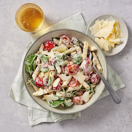 Krämig pastasallad med penne, babyspenat, körsbärstomater och parmesanflarn, serverad i en skål med dryck och extra parmesan bredvid.