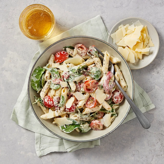 Krämig pastasallad med penne, babyspenat, körsbärstomater och parmesanflarn, serverad i en skål med dryck och extra parmesan bredvid.