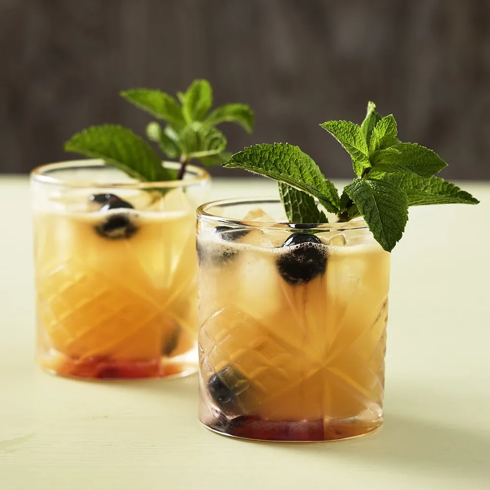 Rosa cocktail i cocktailglas med äppelskiva, jordgubbar och rhubarberstjälkar på bord med shaker i bakgrunden.